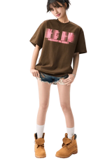 Young woman wearing brown MEDM Phantom Contrast Tee with pink logo, denim shorts, white socks, and tan boots