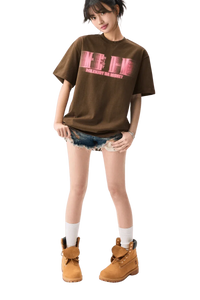 Young woman wearing brown MEDM Phantom Contrast Tee with pink logo, denim shorts, white socks, and tan boots