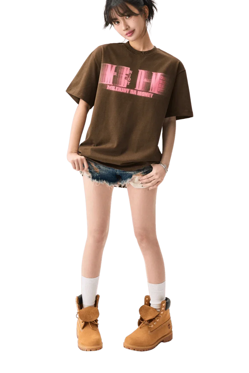 Young woman wearing brown MEDM Phantom Contrast Tee with pink logo, denim shorts, white socks, and tan boots