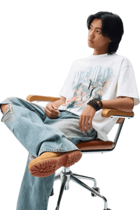 Model wearing a MEDM SS24 Phoenix Series Print Tee with ripped jeans, seated in a modern chair.