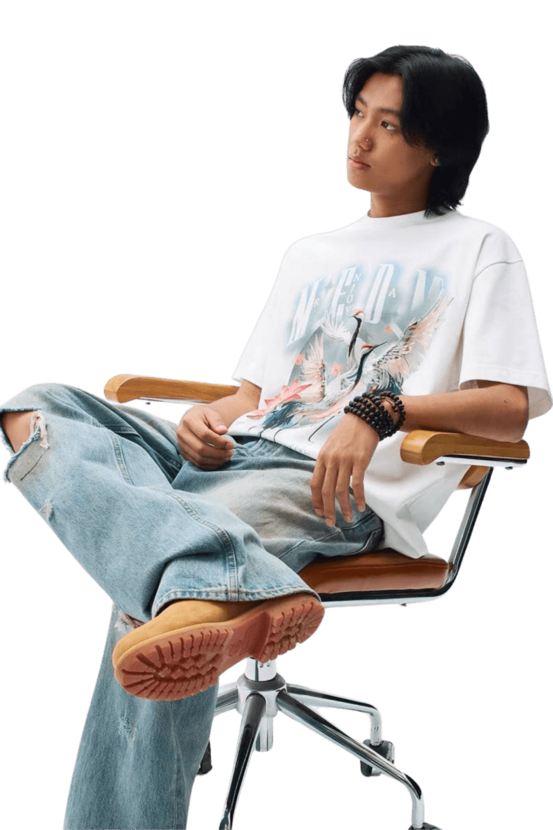 Model wearing a MEDM SS24 Phoenix Series Print Tee with ripped jeans, seated in a modern chair.