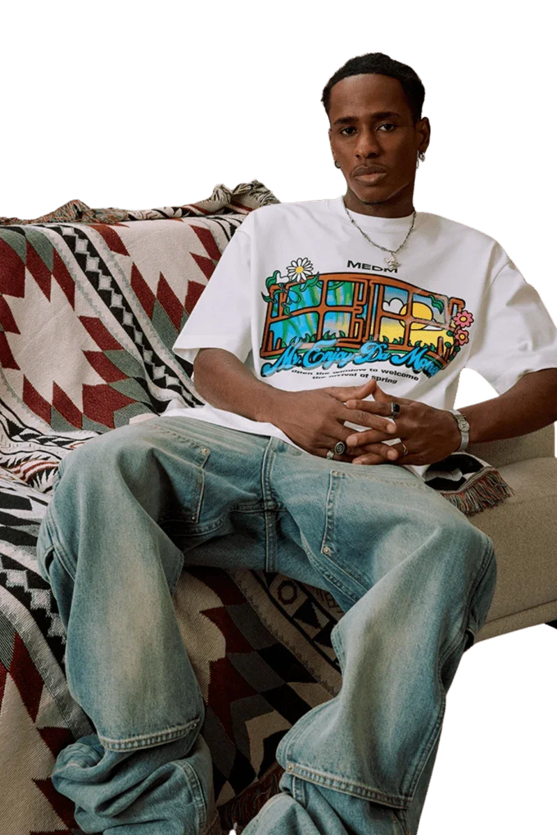 Man wearing white MEDM Window Spring Tee with colorful graphic design sitting on patterned blanket