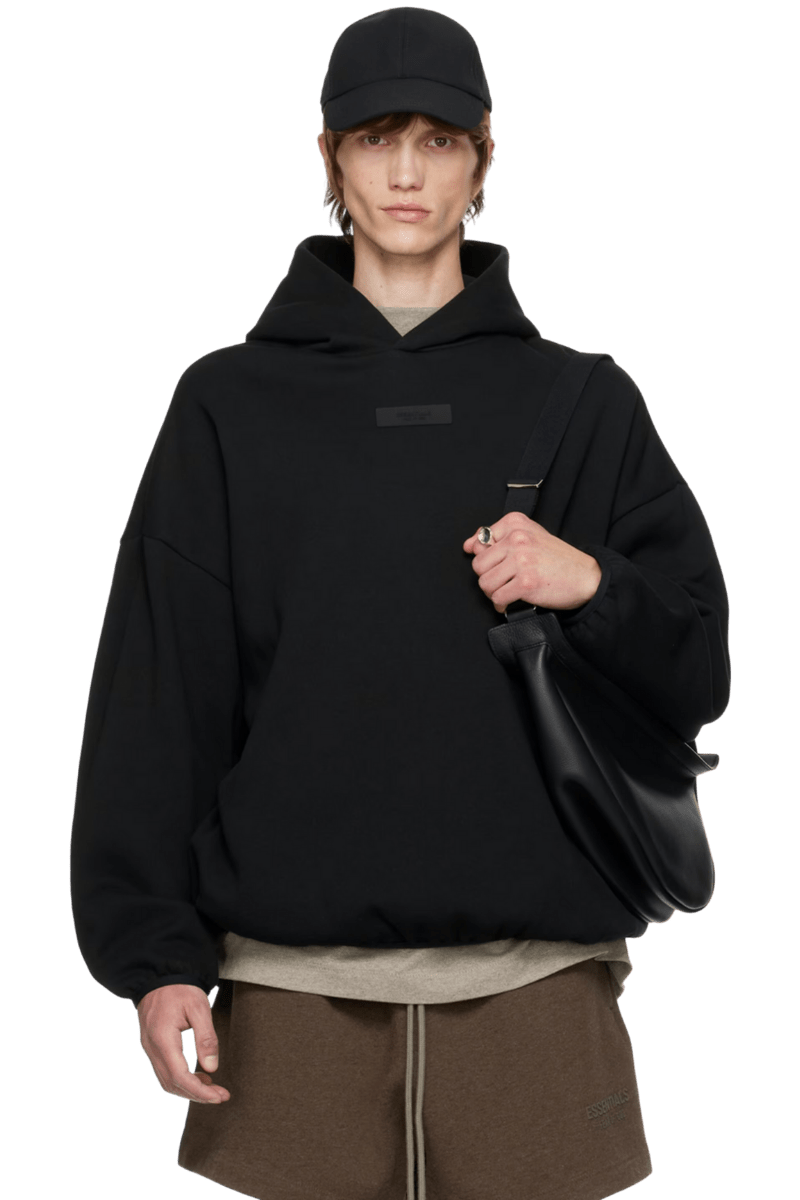 Fear Of God Essentials SS24 Heavy Jersey Arch Logo Hoodie in black, worn with a cap and stylish bag